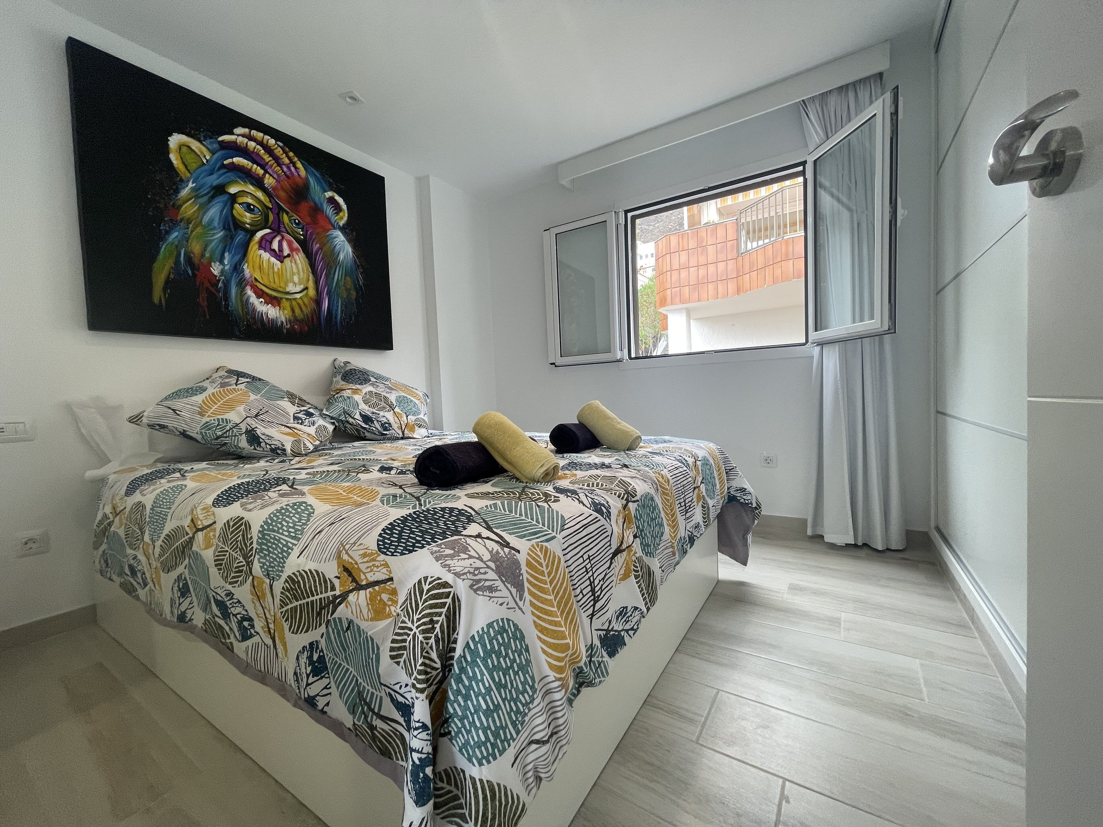 Een slaapkamer met een bed, een kleurrijk schilderij, een raam met gordijnen en uitzicht op gebouwen.