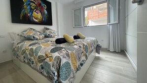 A bedroom with a bed, a colorful painting, a window with curtains, and a view of buildings outside. - Tamara Alto (Acantilados de Los Gigantes)