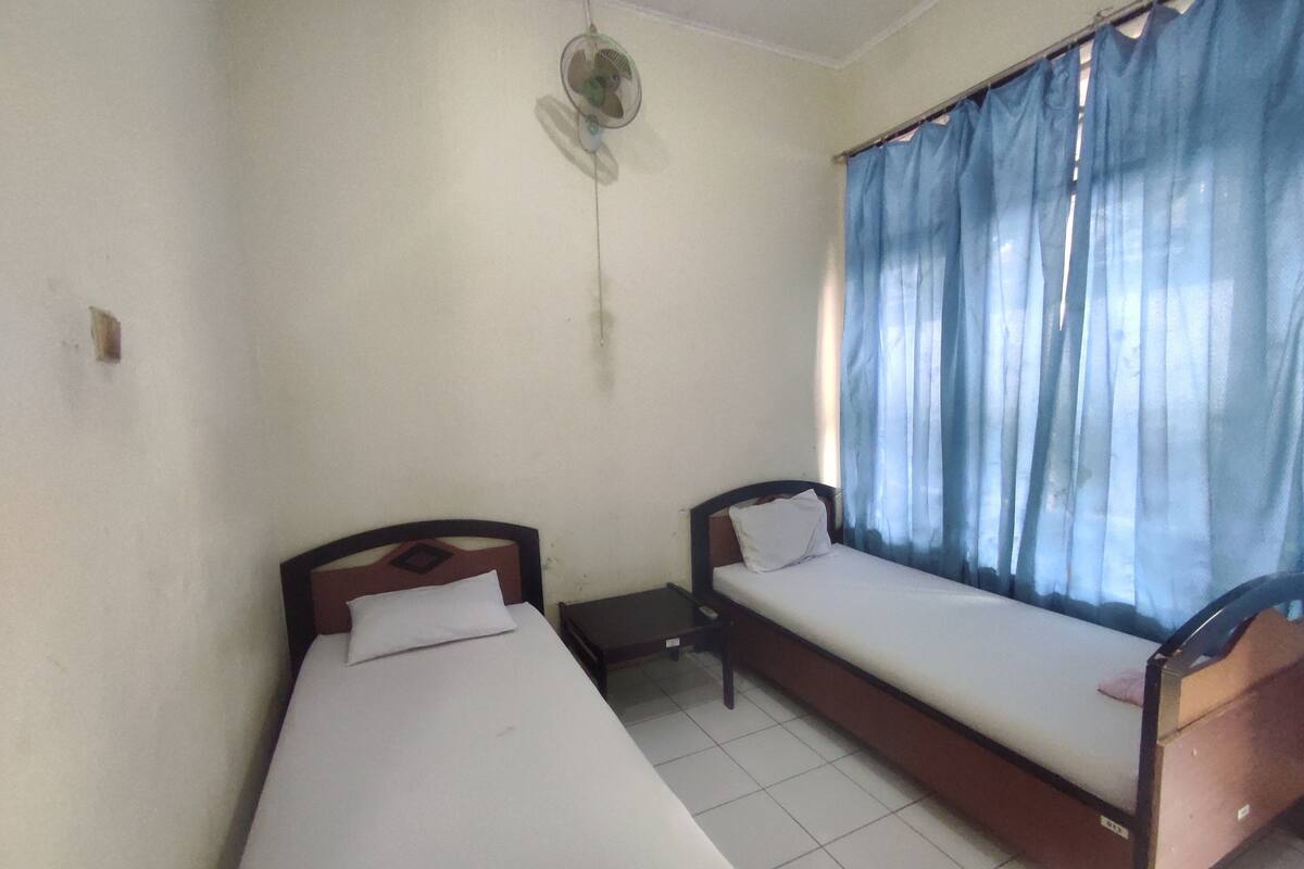 A room with two single beds, a small table, a wall-mounted fan, and a window with blue curtains.