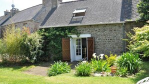A stone house with a sloped roof, a small garden with various plants, and a wooden door. - Gite Coriosolites (Quemper-Guézennec)
