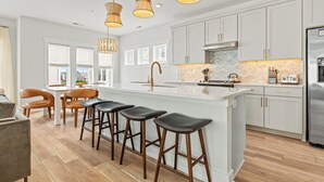 A modern kitchen with a central island, bar stools, pendant lights, and a stainless steel refrigerator. - Seashell Serenity Private Pool + Resort Community (Kill Devil Hills)