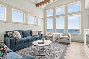 Un salon spacieux avec un canapé bleu, deux fauteuils, une table basse et un tapis à motifs. De grandes fenêtres offrent une vue sur la mer.