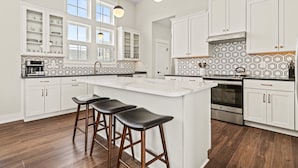 Una cocina moderna con armarios blancos, una isla central con taburetes, horno integrado y un salpicadero decorativo.