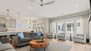 A spacious living room with a grey sectional sofa, two armchairs, a coffee table, and a dining area with a kitchen in the background. - Sunset Soiree - Private Pool + Resort Amenities (Kill Devil Hills)