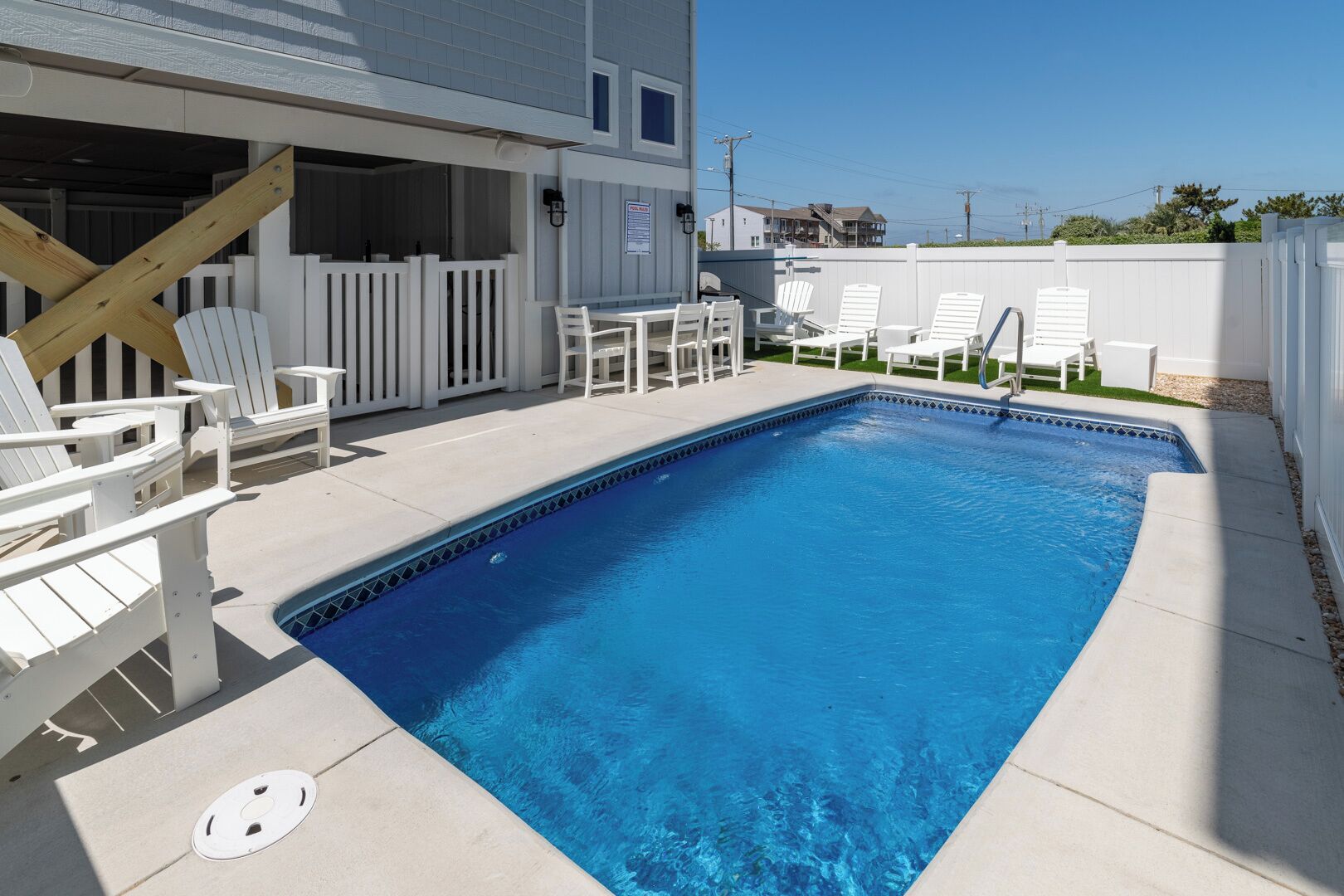A rectangular swimming pool with a tiled edge, surrounded by a concrete deck. There are white Adirondack chairs and a table with chairs on the deck. A wooden deck railing is visible on the left side.