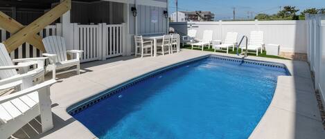 A rectangular swimming pool with a tiled edge, surrounded by a concrete deck. There are white Adirondack chairs and a table with chairs on the deck. A wooden deck railing is visible on the left side.