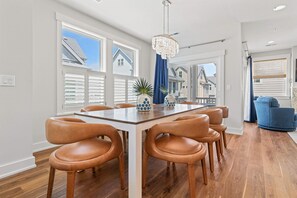 A dining area with a wooden table and leather chairs, large windows, and a view of neighboring houses. - Moonlit Manor - Pets Welcome + Private Pool (Kill Devil Hills)