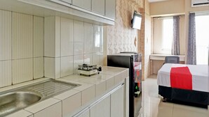 Habitación de hotel con kitchenette, cama con colcha rojo y negro, televisor montado en la pared y ventana con cortinas.