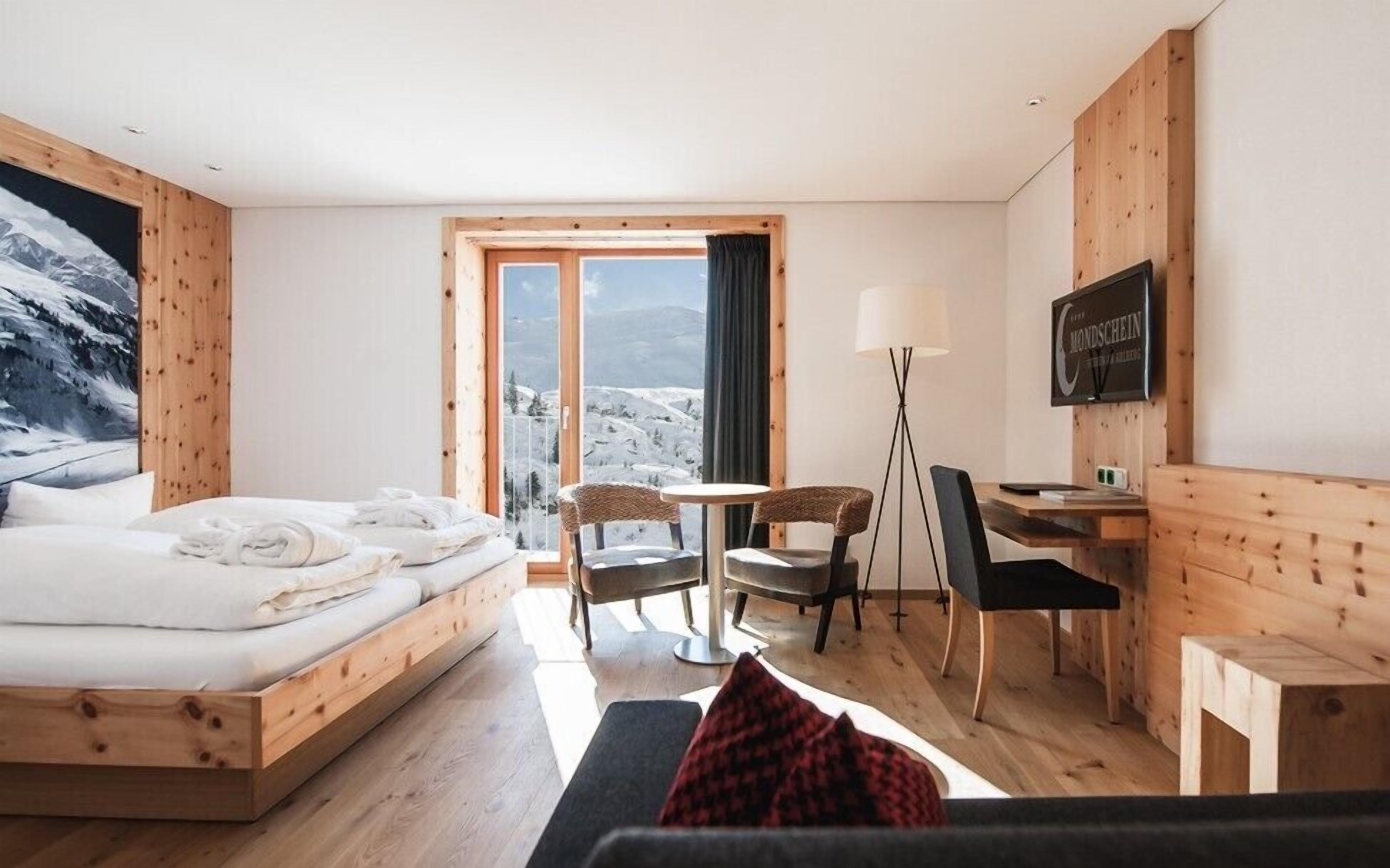 A hotel room with a bed, a desk, two chairs, a TV, and a large window with a mountain view.