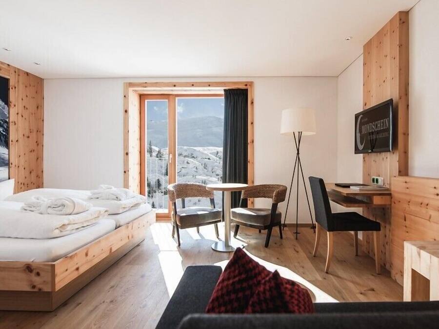 A hotel room with a bed, a desk, two chairs, a TV, and a large window with a mountain view.