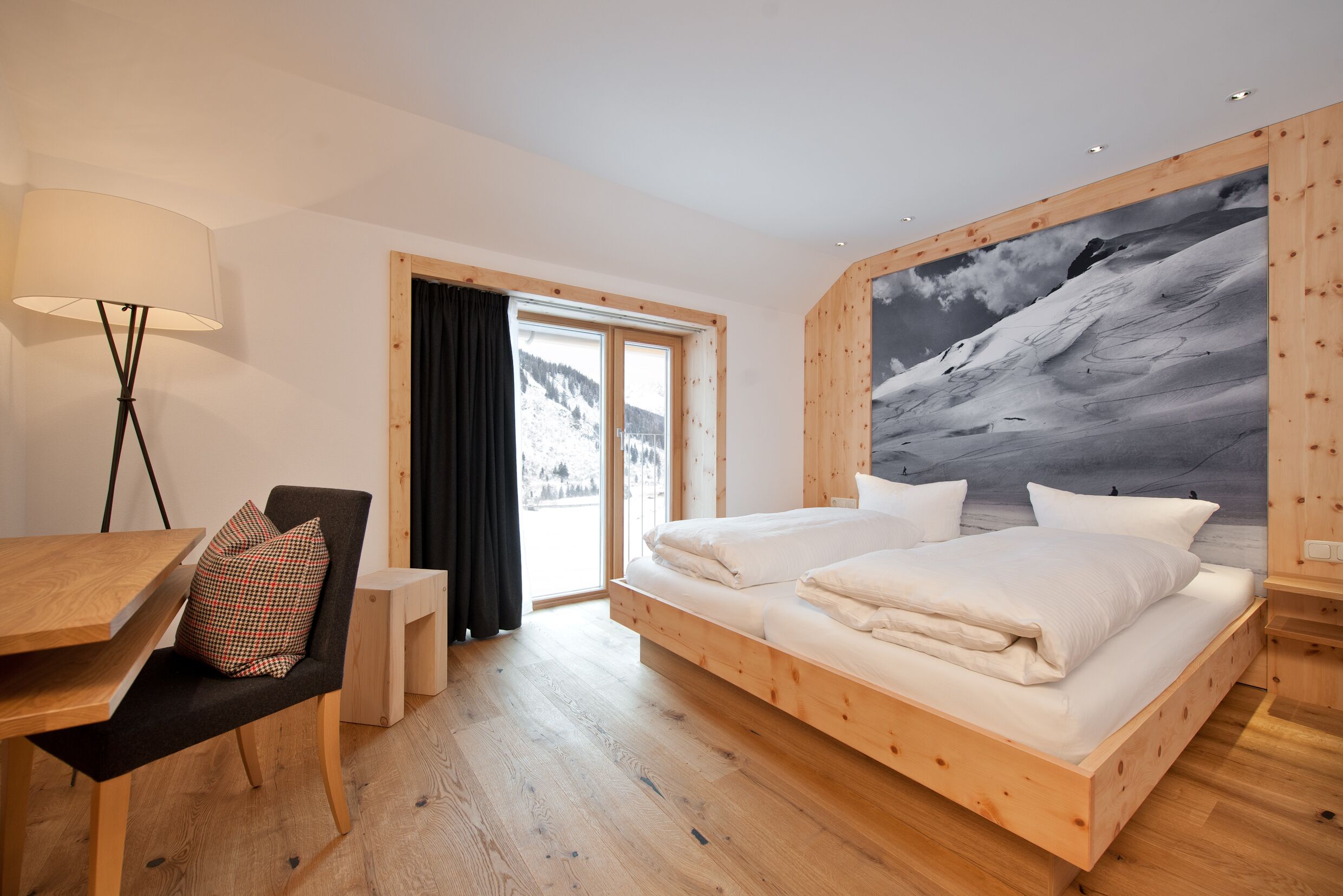 A bedroom with a wooden bed, a desk, a chair, a lamp, and a large window with a view of a snowy mountain.