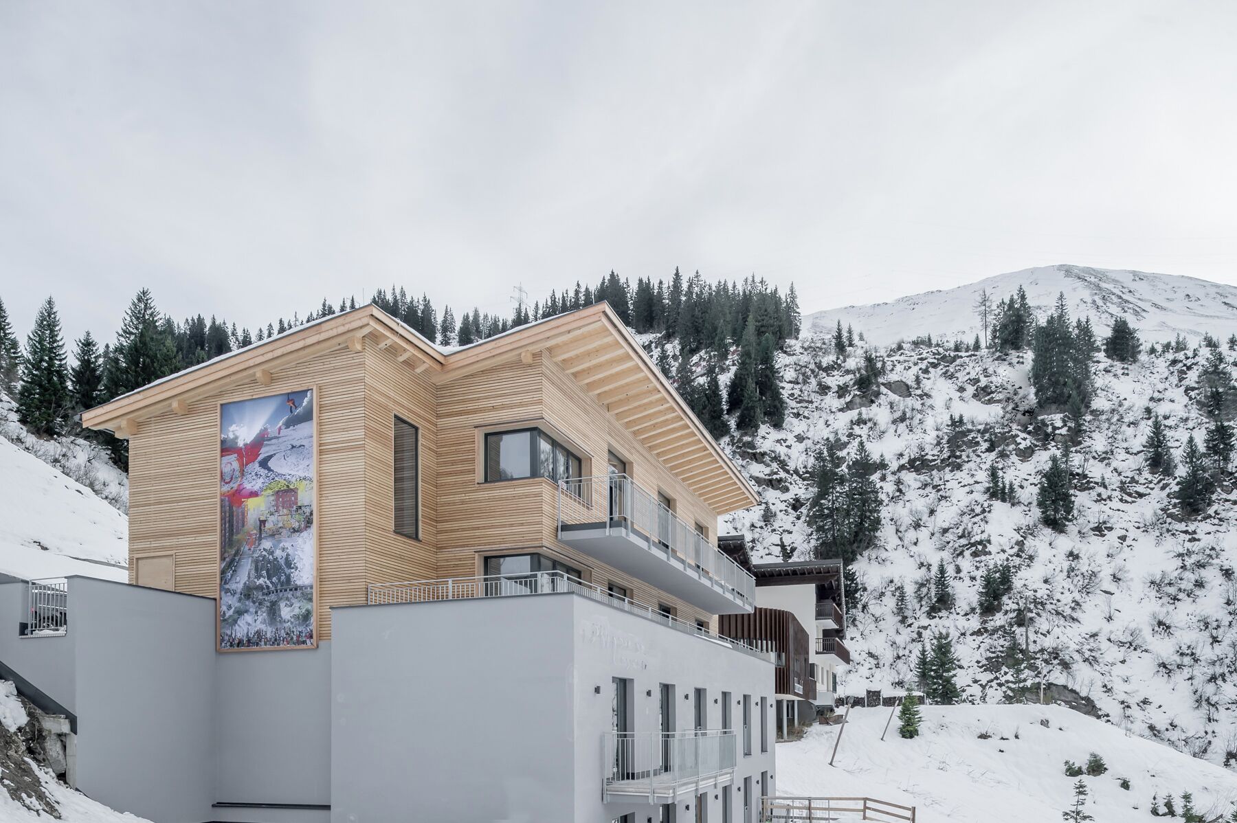 Un edificio moderno con fachada de madera y varios balcones, rodeado de un paisaje montañoso nevado.
