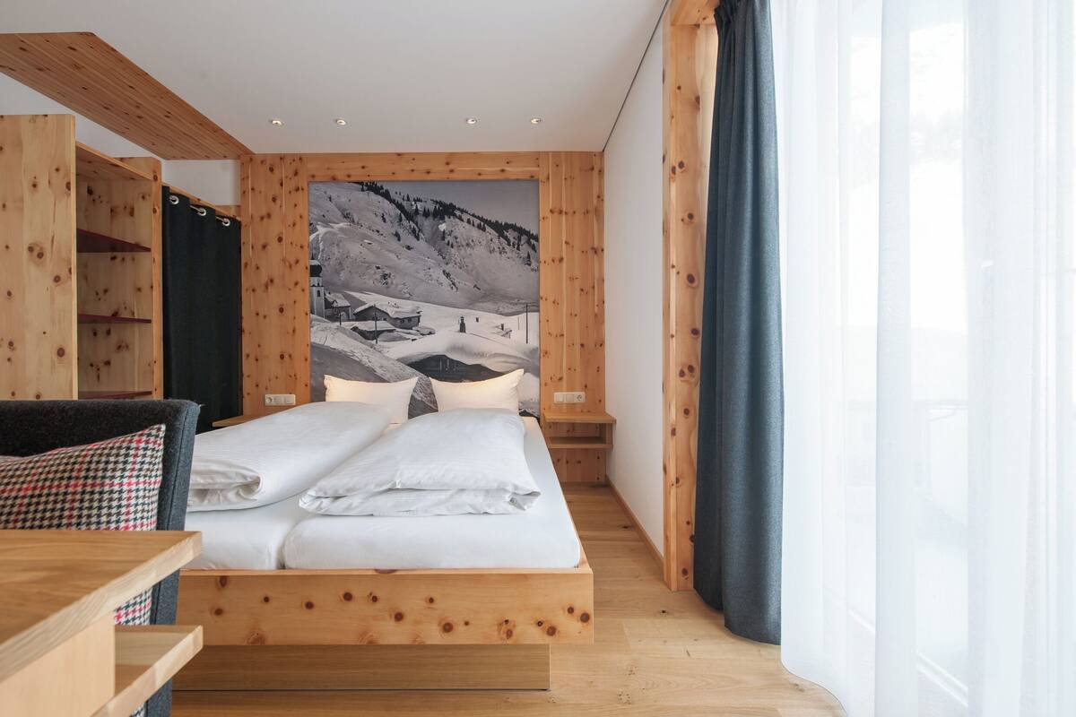 Un dormitorio moderno con una cama de madera, un ventanal con cortinas traslúcidas y un mural con paisaje de montaña en la pared.