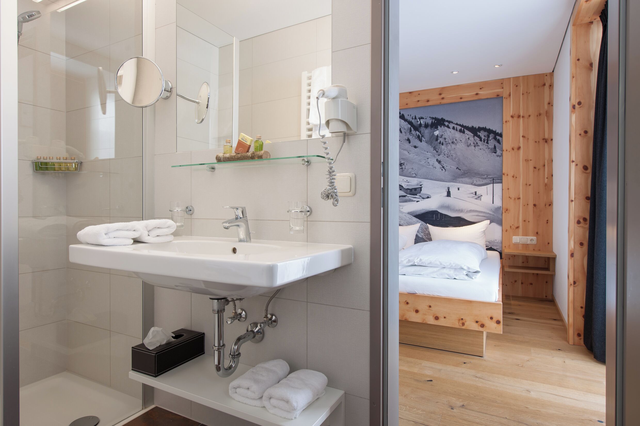 A modern bathroom with a large mirror, sink, and shower. Adjacent is a bedroom with a wooden bed and a mountain view.