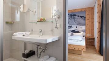 A modern bathroom with a large mirror, sink, and shower. Adjacent is a bedroom with a wooden bed and a mountain view.