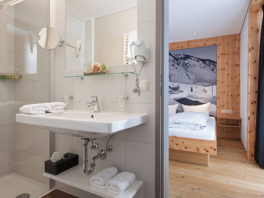 A modern bathroom with a large mirror, sink, and shower. Adjacent is a bedroom with a wooden bed and a mountain view.