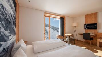 Ein Hotelzimmer mit zwei Betten, einem Schreibtisch, einem Sessel, einem Fernseher und Blick auf einen verschneiten Berg.