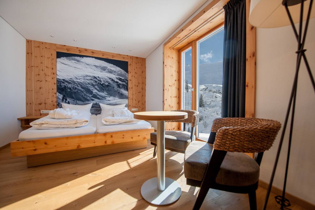 Un dormitorio con una cama de madera, una mesa redonda, una silla y un ventanal con vistas a montañas.