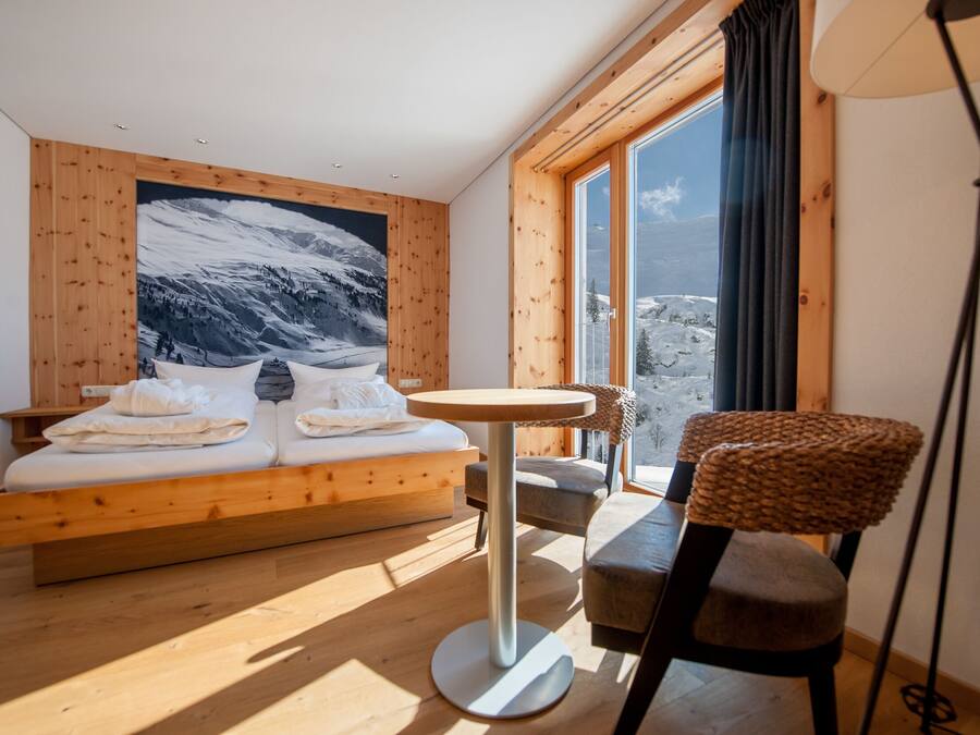 Un dormitorio con una cama de madera, una mesa redonda, una silla y un ventanal con vistas a montañas.