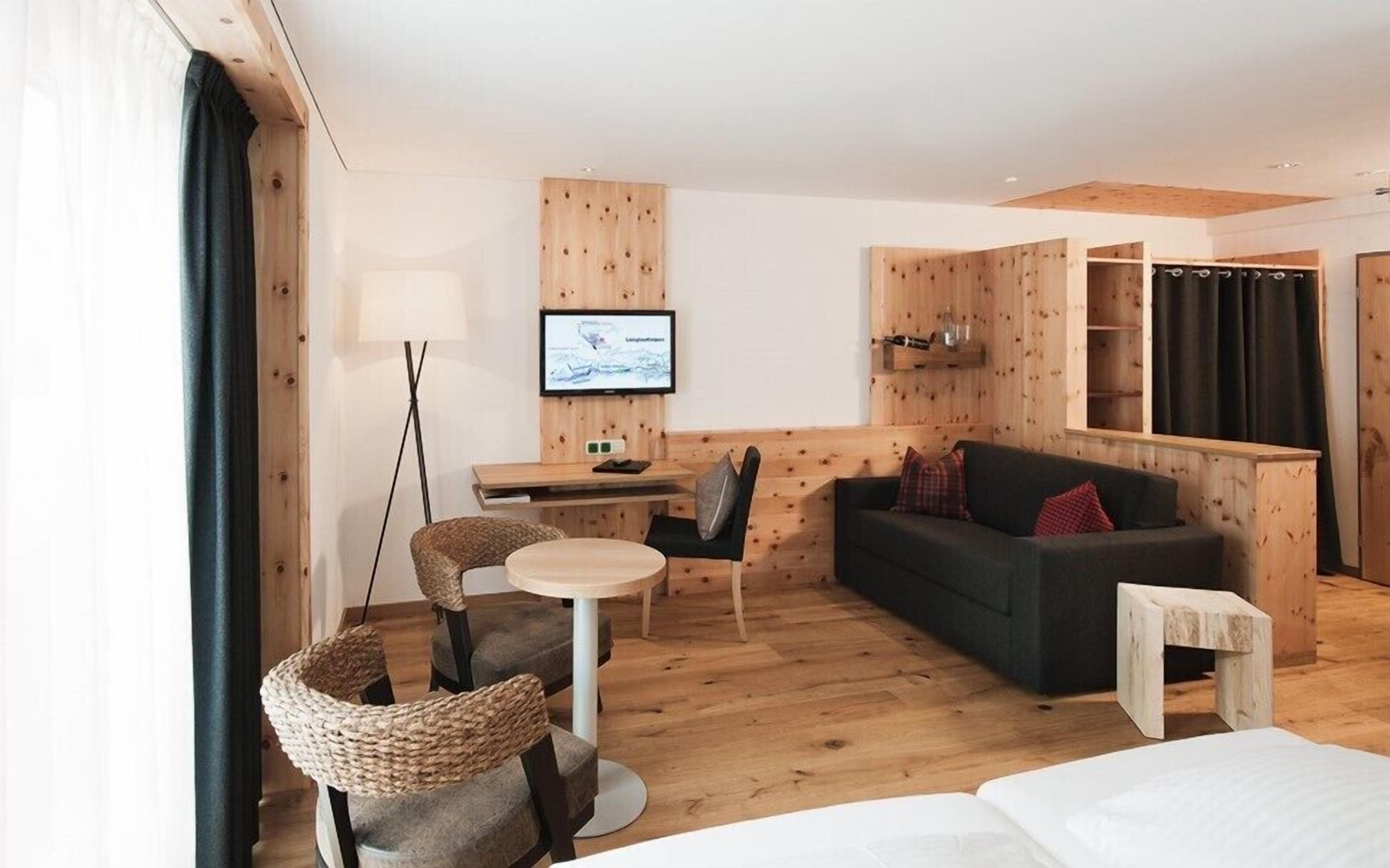 A modern hotel room with a wooden floor, a sofa, a small table, a chair, a TV, and a bookshelf.