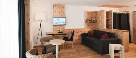 A modern hotel room with a wooden floor, a sofa, a small table, a chair, a TV, and a bookshelf.