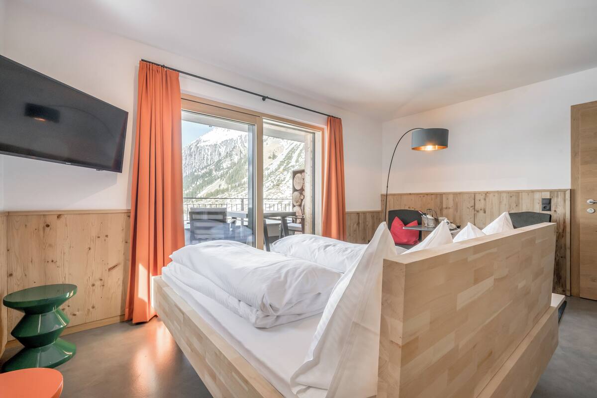 Una habitación de hotel moderna con una cama de madera, un televisor de pantalla plana, un escritorio de madera y vistas a montañas desde la ventana.