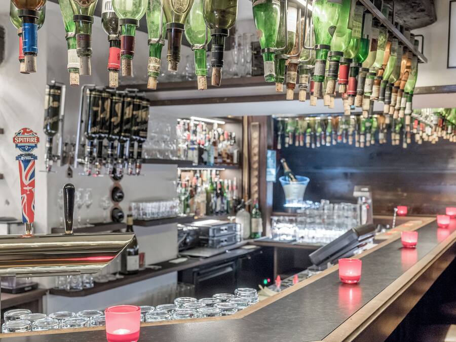 A bar area with multiple liquor bottles hanging from the ceiling, a Spitfire beer tap, and a bar counter with glasses and a red candle.