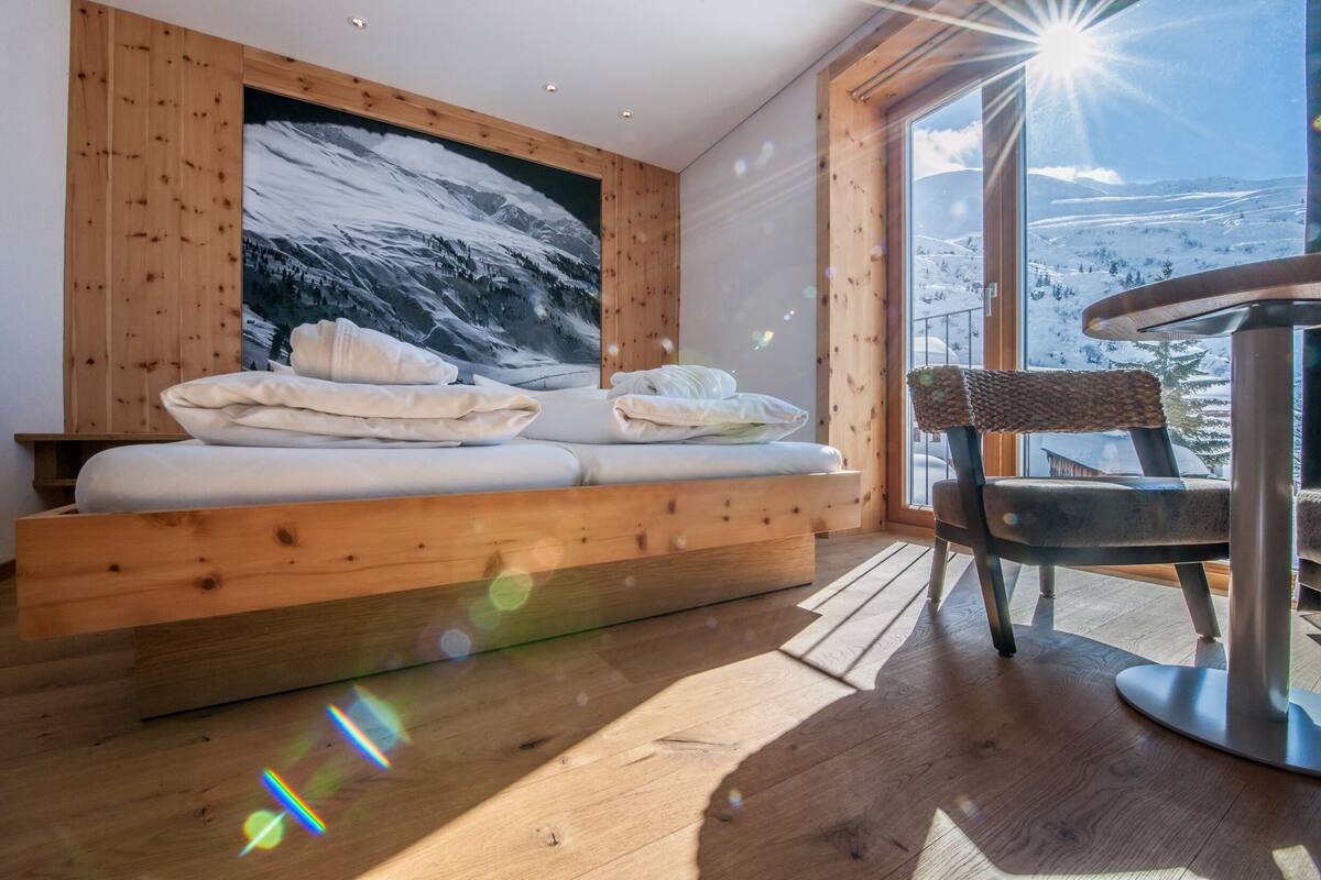 Una habitación con pared de madera, un ventanal grande, una cama con ropa blanca y una silla de mimbre.