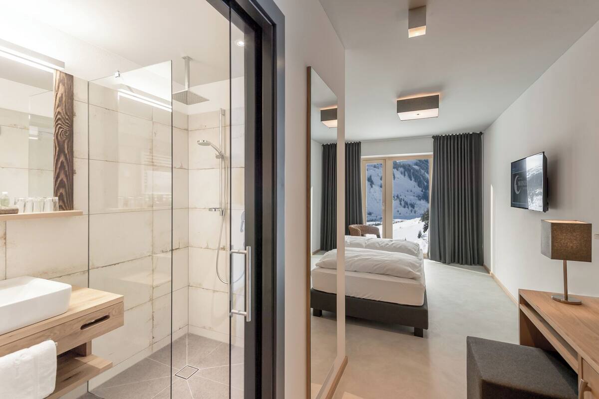 Una habitación de hotel moderna con cabina de ducha de vidrio, una cama grande, un televisor y vistas a montañas nevadas.