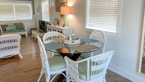 A dining area with a glass-top table surrounded by white wicker chairs, a television, and a lamp on a side table. - 2 Bedroom Bay Front Home - Monthly Rentals (Dauphin Island)