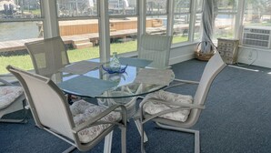 A sunroom with a glass table and chairs, large windows, and a view of a dock and buildings. - 2 Bedroom Bay Front Home - Monthly Rentals (Dauphin Island)