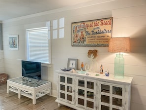 Um quarto com mesa de centro de vidro, uma cadeira de vime branca, um armário branco com portas de vidro e uma parede com um pôster de tema praiano.