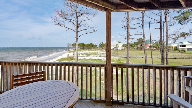 A wooden deck with a table and chairs overlooking a beach with a fence and trees. - Gulf-view condo steps away from beach! 2 Community Pools!  (Dauphin Island)