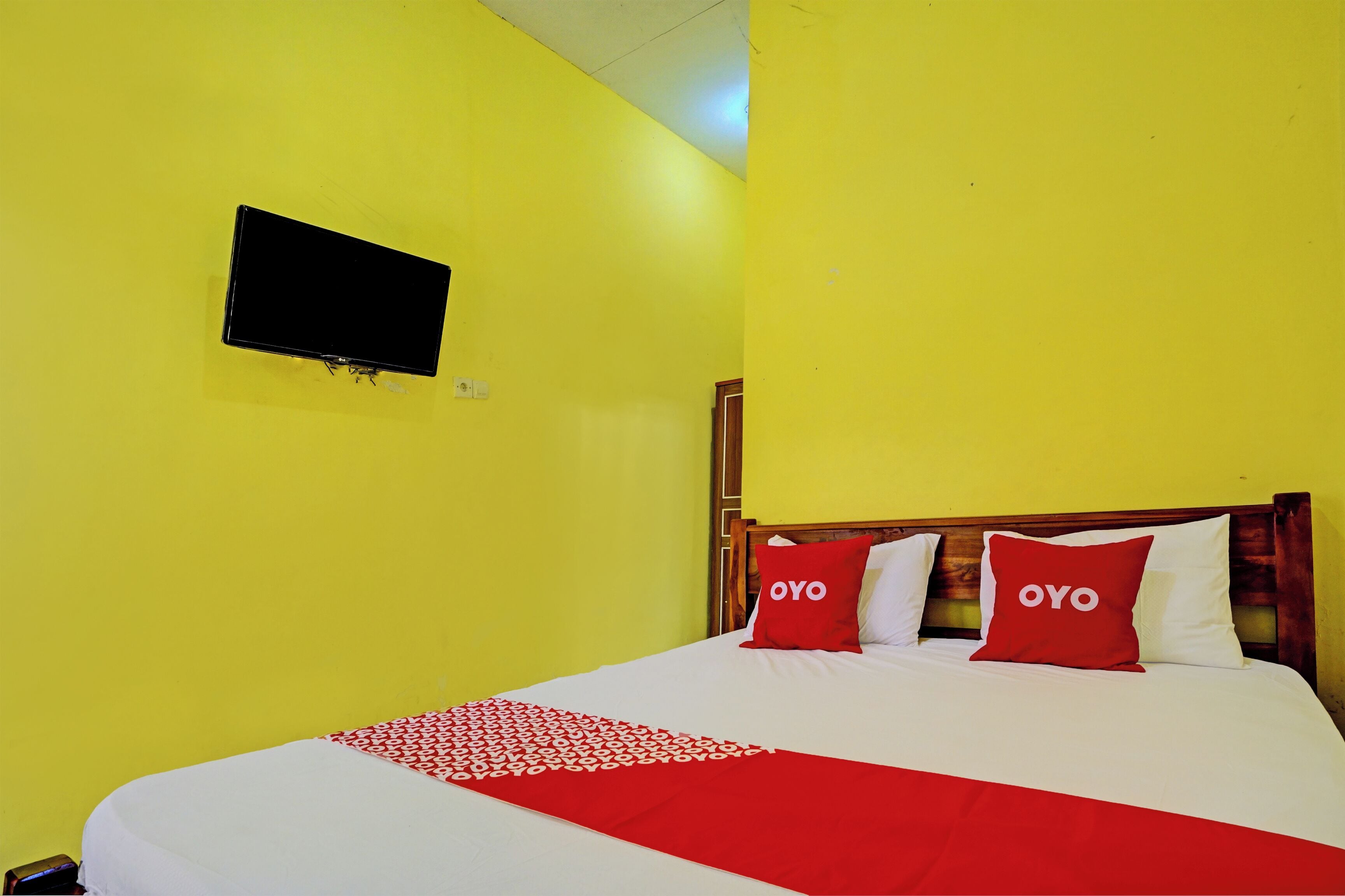 A hotel room with a bed, two red pillows with 'OYO' branding, a flat-screen TV mounted on the wall, and a red and white patterned throw blanket.