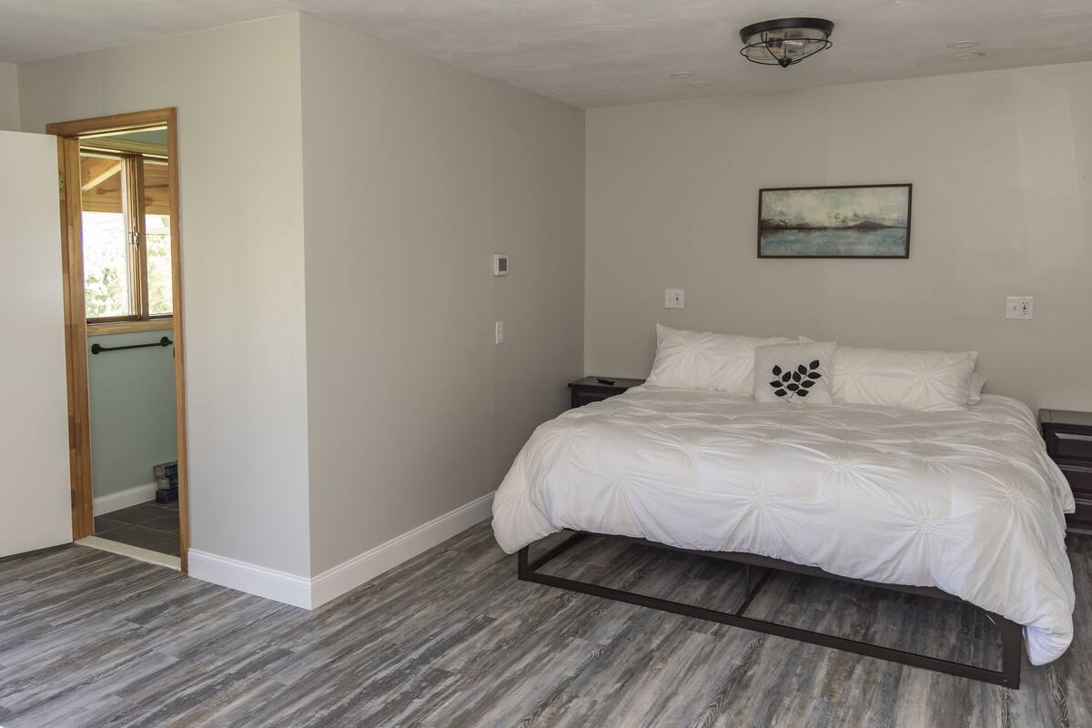 A bedroom with a bed, a nightstand, a wall-mounted painting, and a wooden-framed mirror.