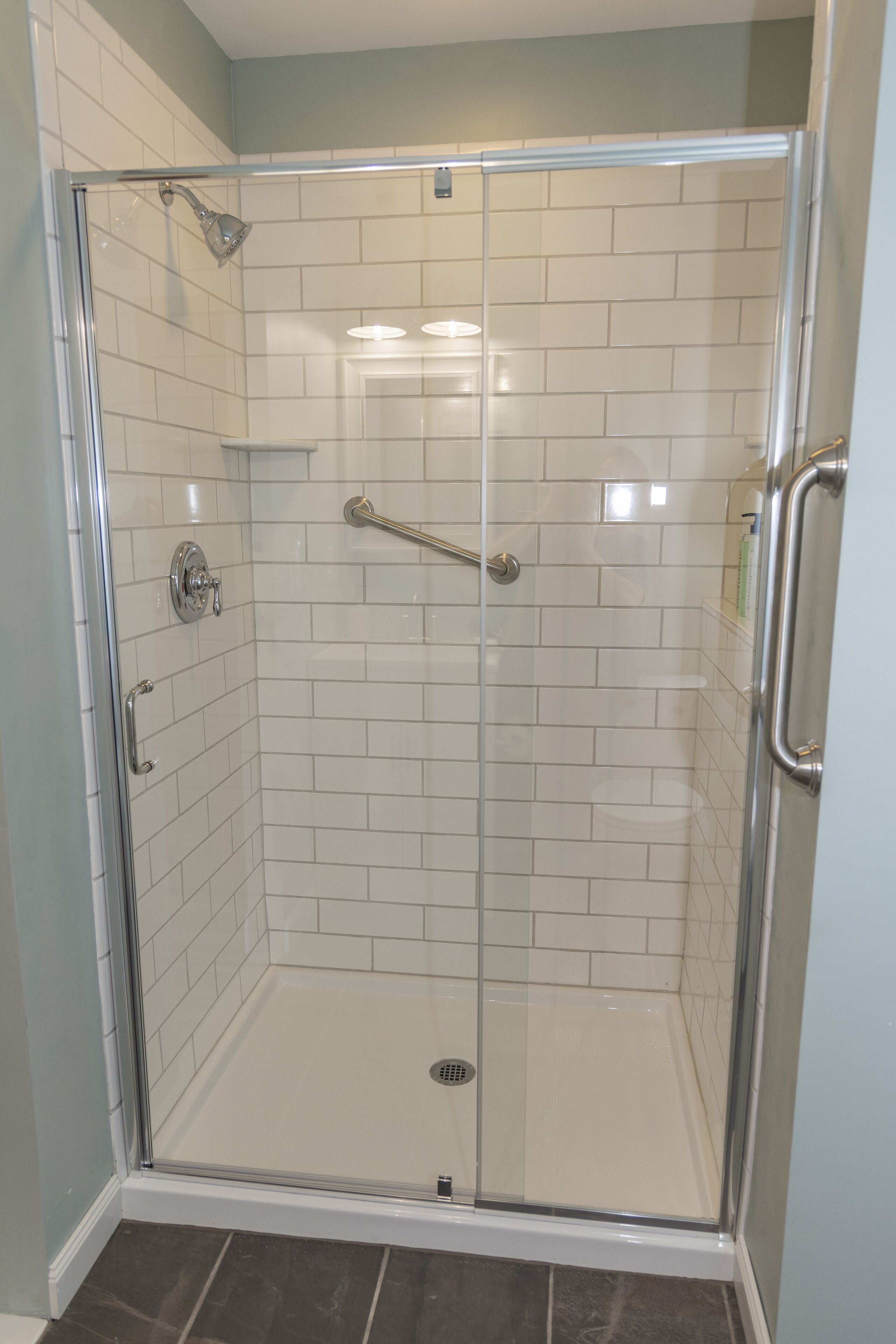 Une cabine de douche fermée par des parois en verre, avec des murs recouverts de carreaux blancs et une poignée en métal.