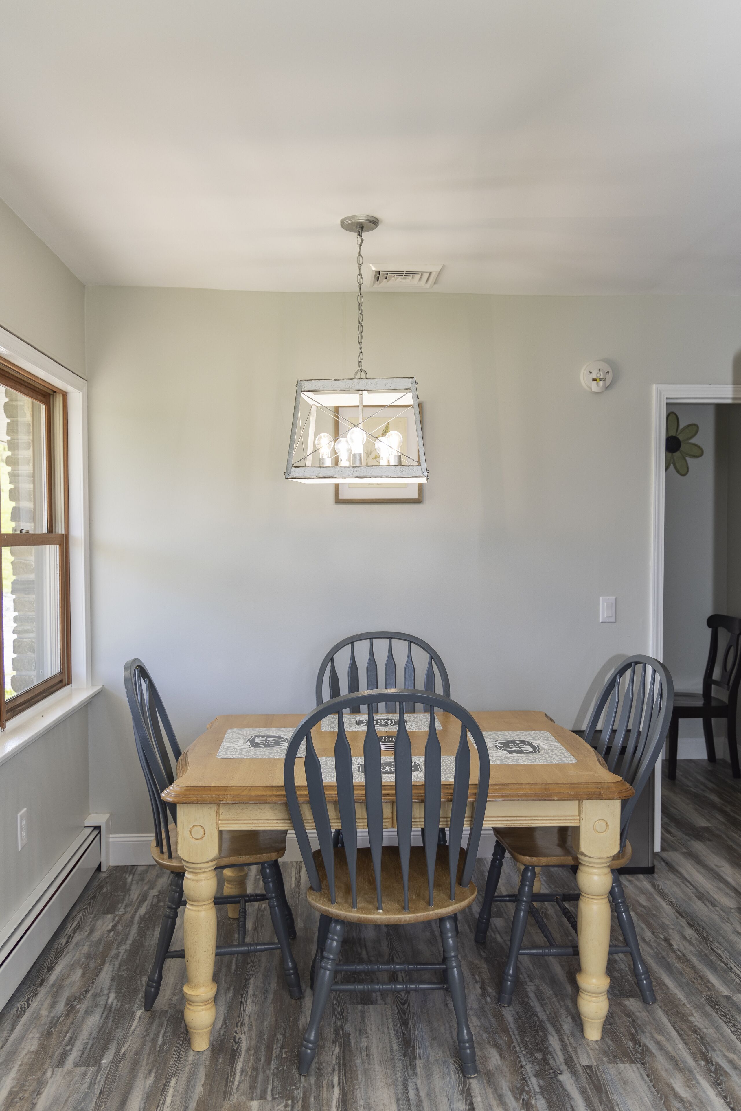 A dining room with a wooden table, four chairs, a hanging light fixture, and a window with blinds.