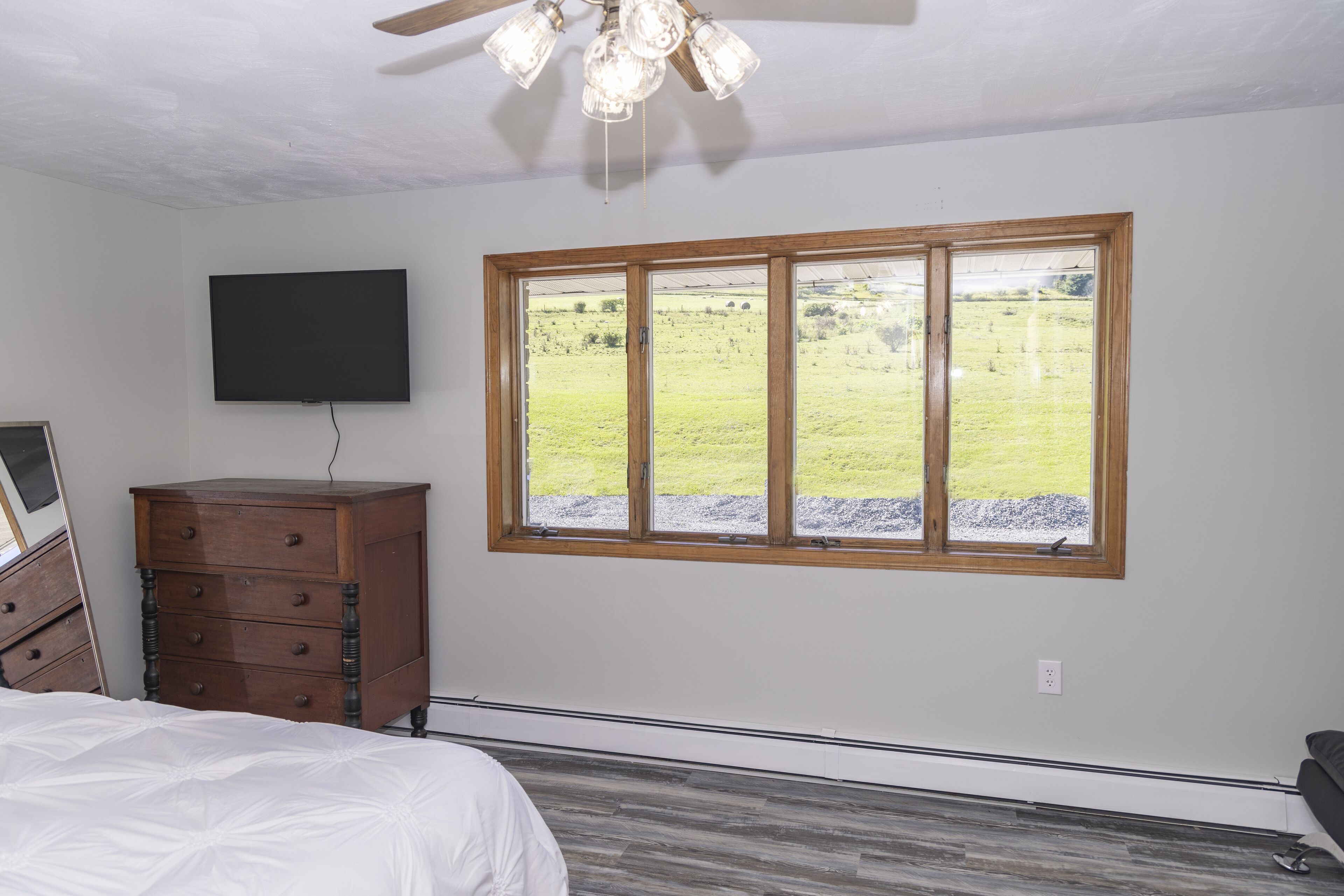 A bedroom with a wooden dresser, a flat-screen TV, a large window with a view of a grassy field, and a ceiling fan.