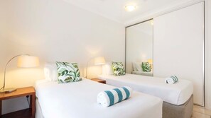 Two single beds with green leaf-patterned pillows and bedside tables with lamps, set against a neutral backdrop.