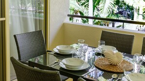 Una mesa de comedor dispuesta con platos blancos, vasos transparentes y un cuenco decorativo, rodeada de sillas de mimbre, con vista a follaje tropical a través de una ventana.