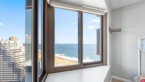 Een kamer met een groot raam met uitzicht op een strand en de oceaan, met hoogbouw op de achtergrond.