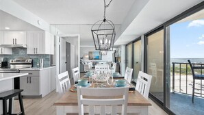 Een moderne keuken met witte kasten, een eethoek met een houten tafel en witte stoelen, en een balkon met uitzicht op zee.