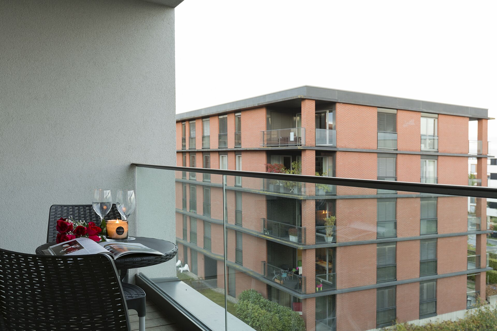 A balcony with a table set for two, a candle, and a magazine. The balcony overlooks a modern brick building with multiple balconies.