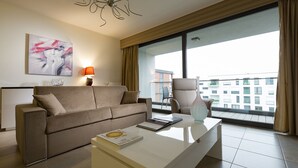 A modern hotel room with a sofa, a coffee table, a lamp, and a view of the city through large windows. - Villa Doria  (Luxembourg City)