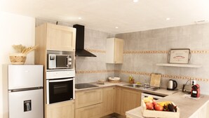 Een moderne keuken met houten kastjes, een ingebouwde magnetron, oven en een zwarte afzuigkap. Op het aanrecht staat een mand met fruit en aan de muur hangt een lijst.