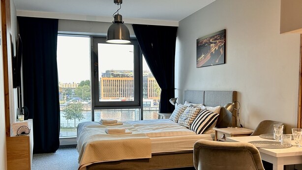 Una habitación de hotel moderna con una cama grande, un escritorio y vistas de la ciudad a través de amplias ventanas.