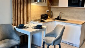 Una cocina compacta con encimera blanca, un microondas Bosch y una cafetera. Hay dos sillas grises y una pequeña mesa de comedor con platos y vasos azules.