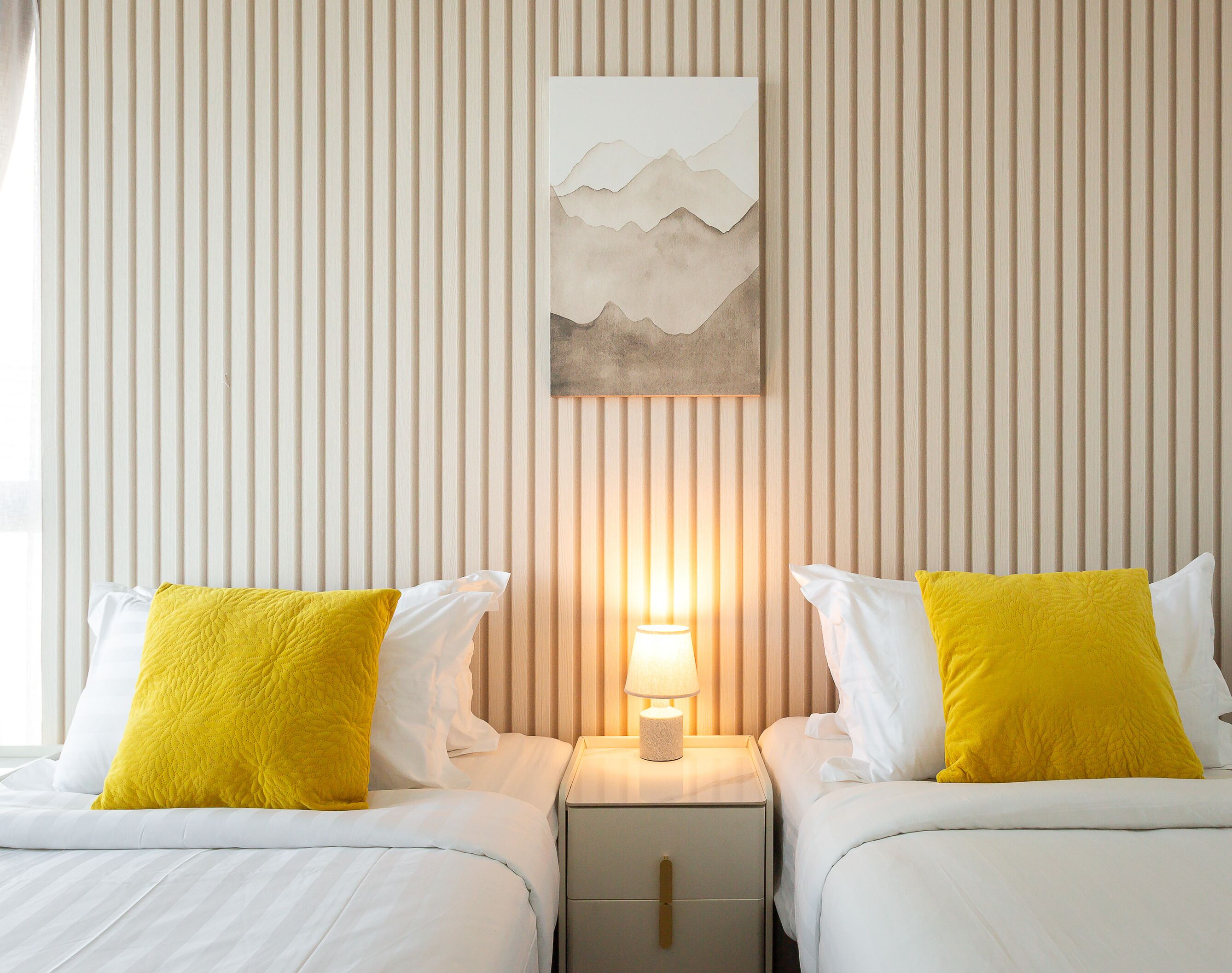 A hotel room with two beds, each with white bedding and a yellow pillow. There is a nightstand with a lamp between the beds.