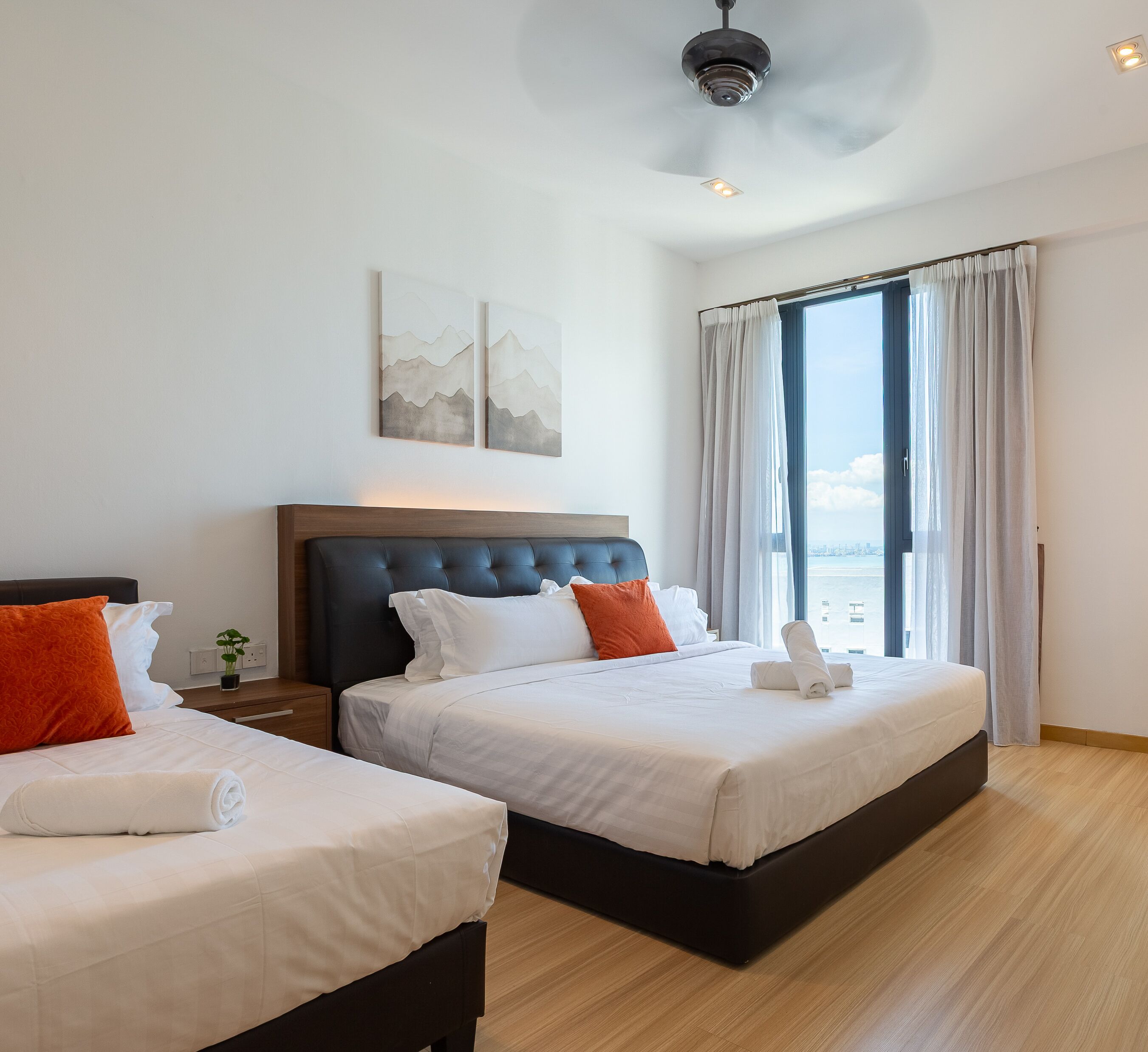 A hotel room with two beds, a large window with curtains, a ceiling fan, and artwork on the wall.
