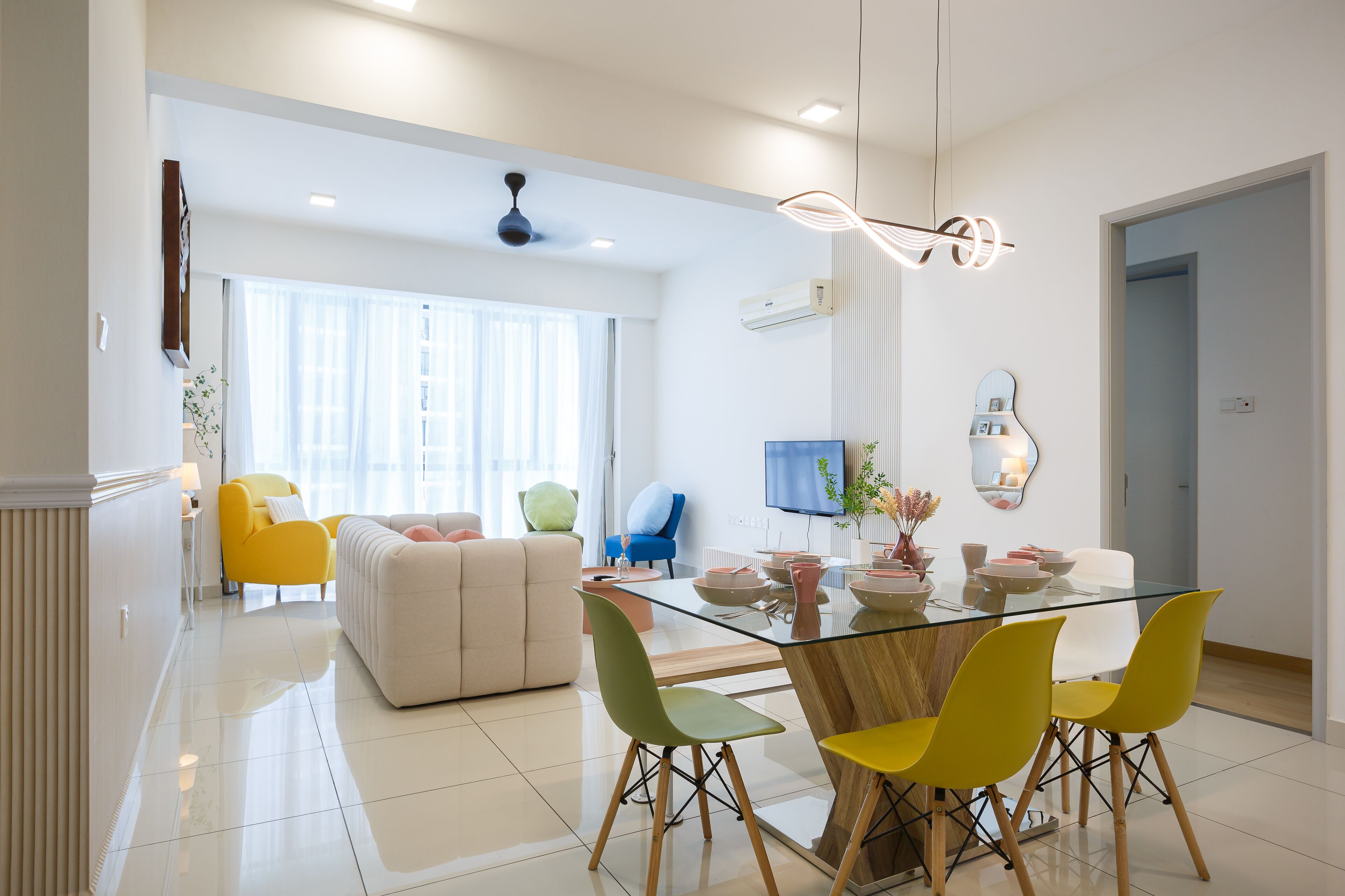 Ein modernes Wohnzimmer mit einem Glastisch, gelben und grünen Stühlen, einem beigen Sofa und einem blauen Sessel.